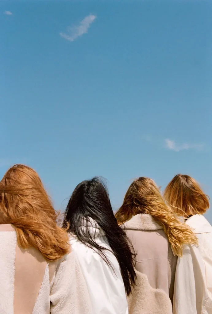Four women with flowing hair stand together, gazing at a vast blue sky