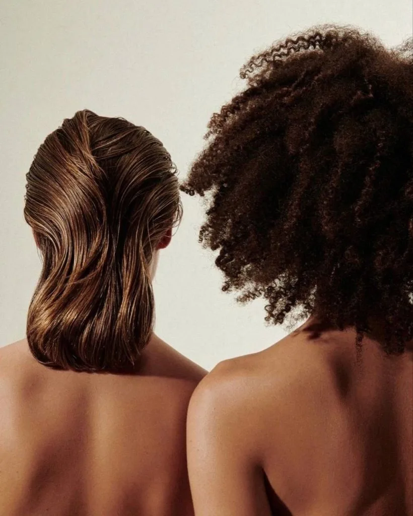 Two individuals stand closely together, showcasing contrasting hairstyles against a neutral background