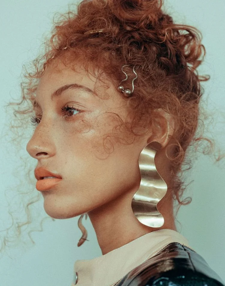 A woman with curly hair displays an artistic metallic earring, posing gracefully