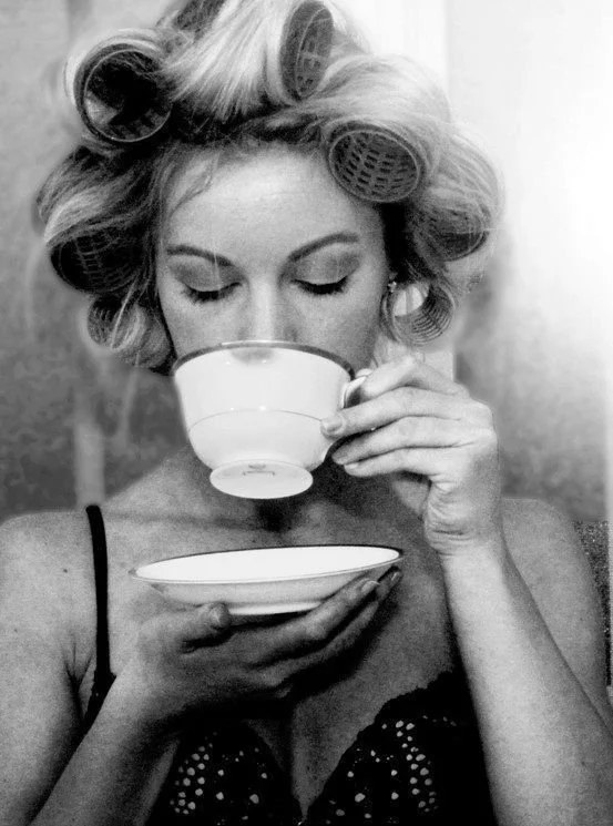 A woman with hair rollers sips tea from a delicate cup and saucer