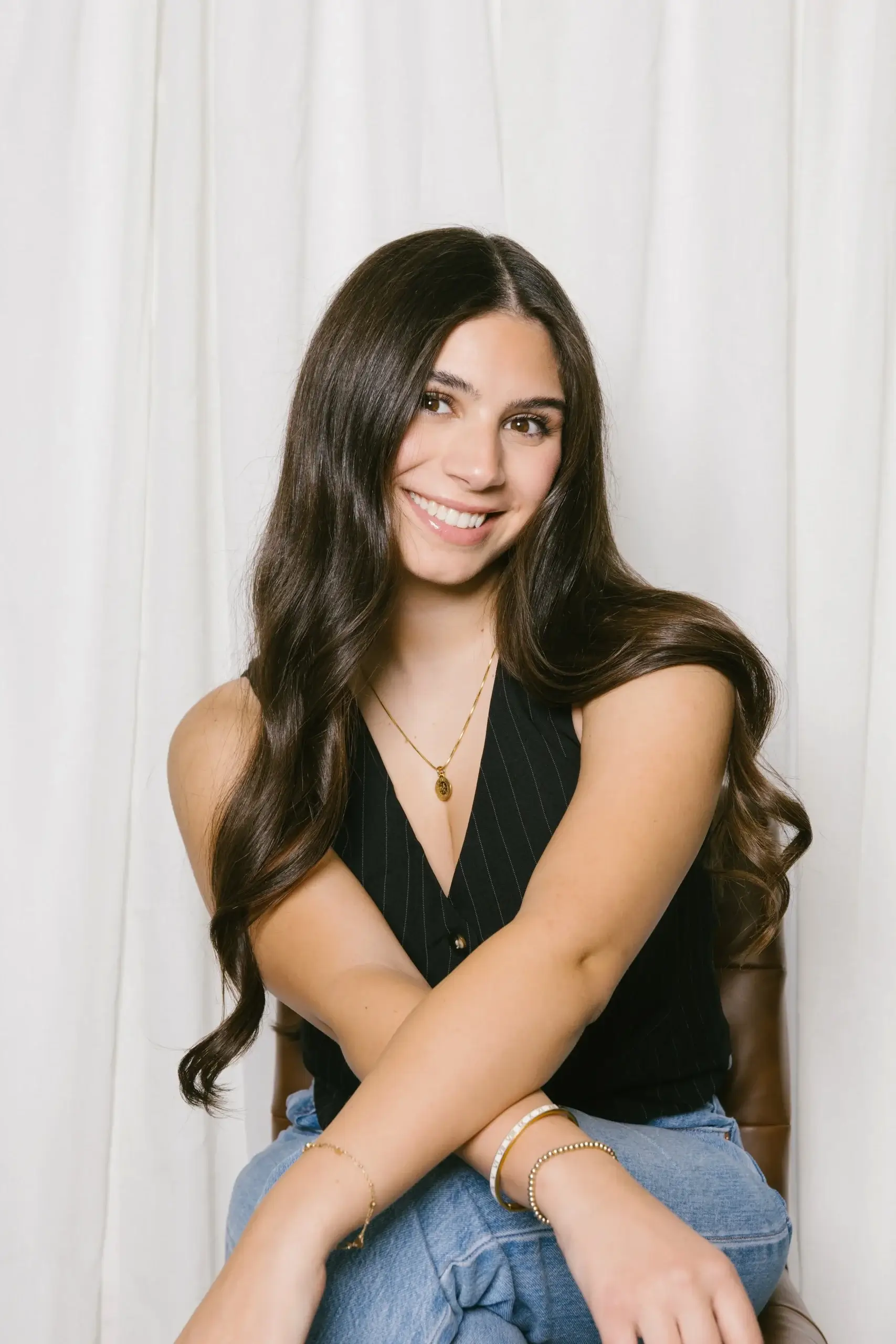 A young woman with long brown hair smiles while sitting against a light backdrop