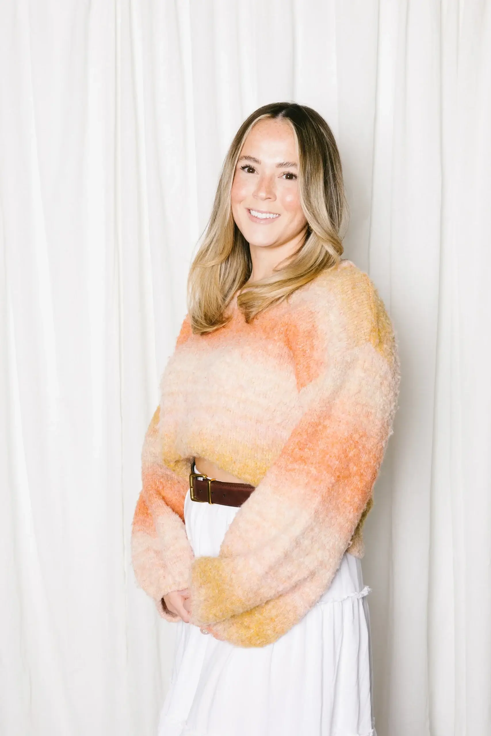 A person stands smiling in a colorful, textured sweater against a white backdrop