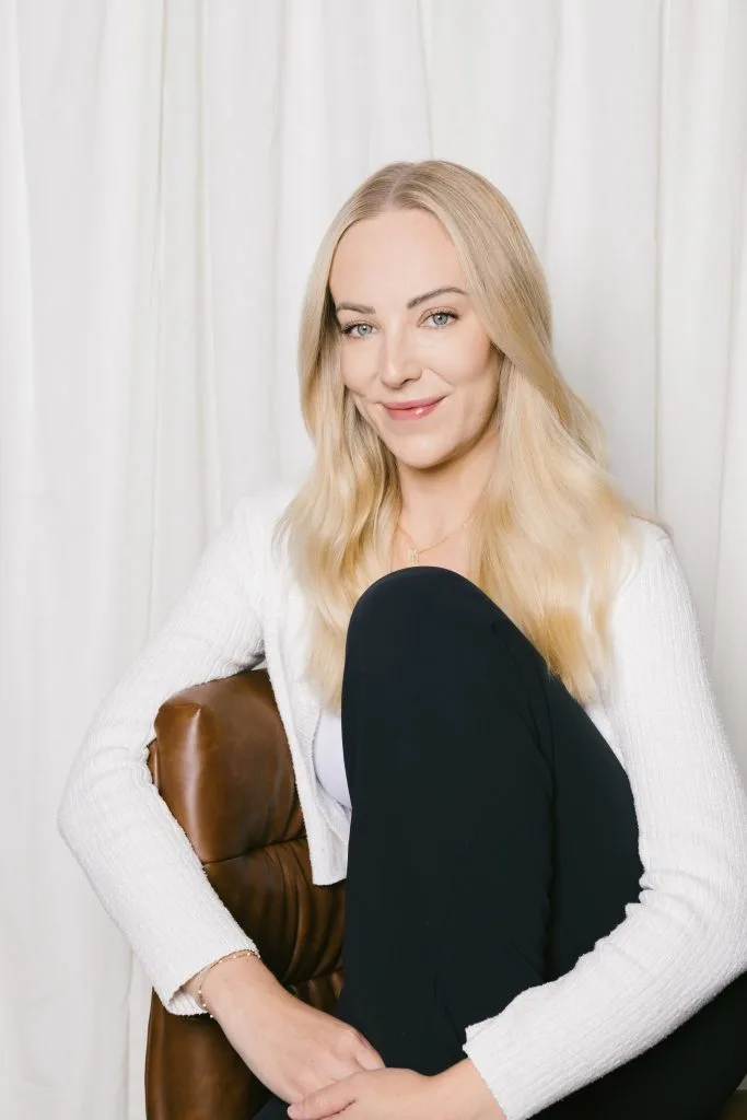 A woman with long blonde hair sits comfortably, smiling while resting her chin on her knee