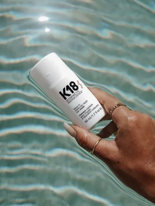A hand holds a bottle of K18 hair product above shimmering water