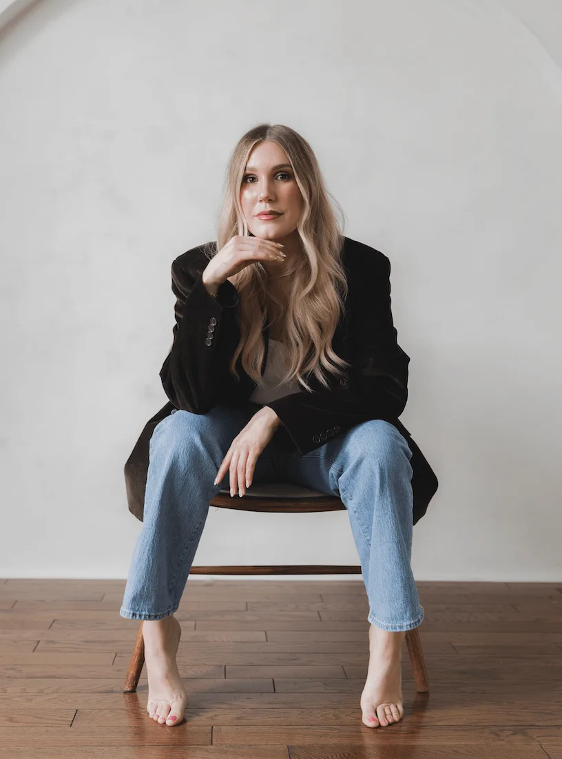 A woman with long hair sits casually on a wooden chair, barefoot and thoughtful