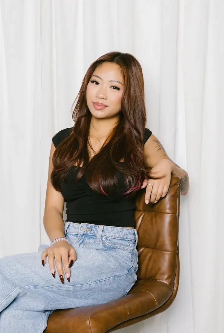 A woman with long hair sits confidently on a chair, wearing casual attire