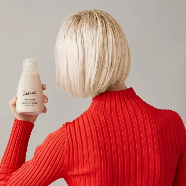A person with a bob haircut holds a bottle of hair product, wearing a red ribbed sweater
