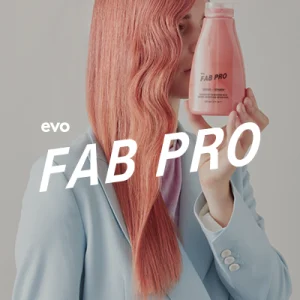 A person with long, wavy pink hair holds a shampoo bottle close to their face