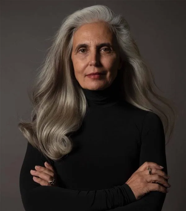 A confident woman with long, flowing gray hair stands with crossed arms