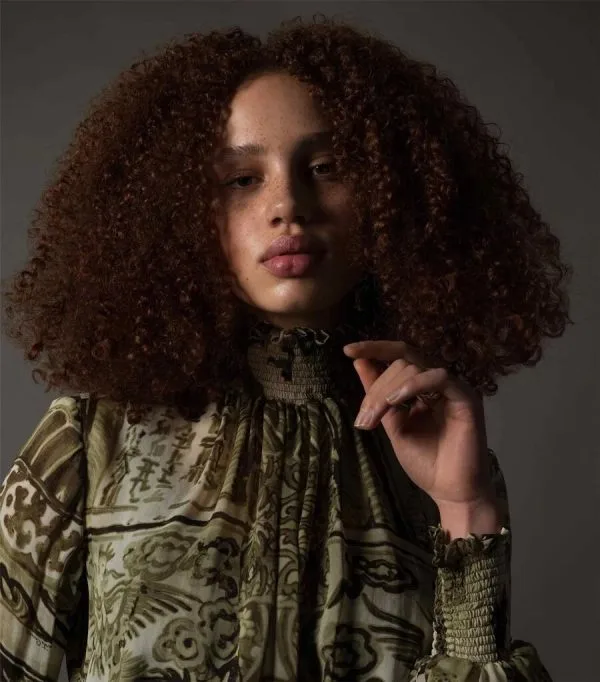 A model with voluminous curly hair poses thoughtfully in elegant attire